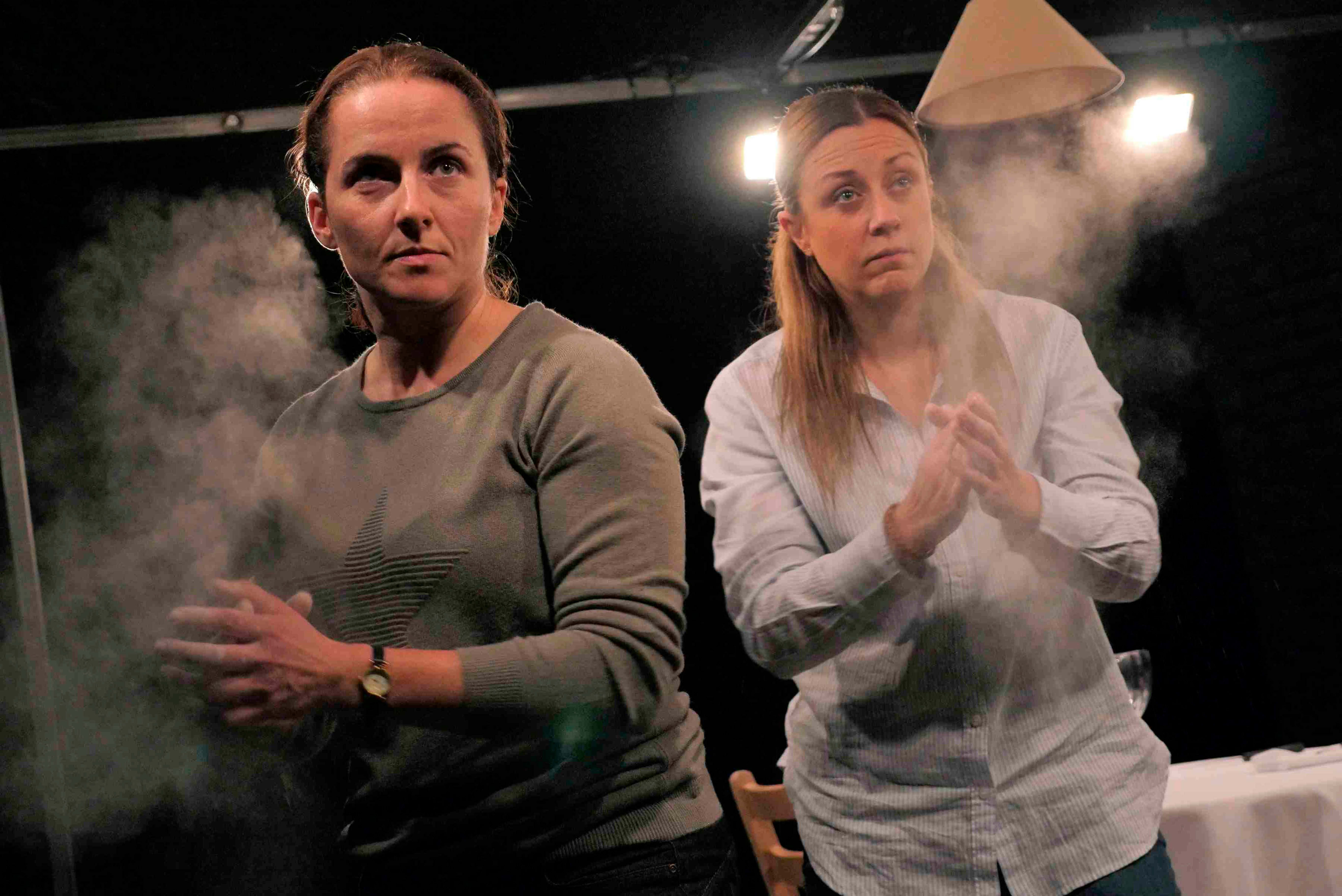 Two women stand on a dimly lit stage, clapping powder from their hands, creating a cloud of dust. One wears a gray sweater with a star design, and the other wears a light button-up shirt. The atmosphere is tense and dramatic. Photo credit Keith Pattison